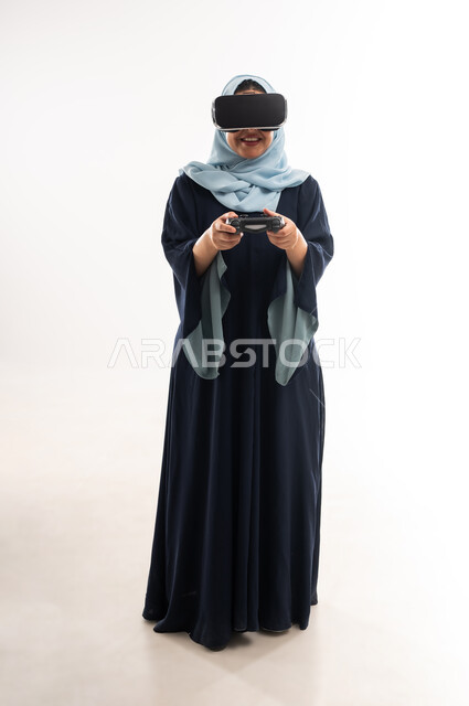 استخدام الأجهزة والتقنيات الحديثة، ايماءات الاستمتاع والاندماج والتفاعل، بورتريه لإمرأة عربية خليجية عمانية مبتسمة ترتدي الحجاب والعباءة تضع نظارة الواقع الإفتراضي وتمسك بيدها عصا التحكم، اللعب بألعاب الفيديو الترفيهية، صورة الجسم بالكامل، خلفية بيضاء