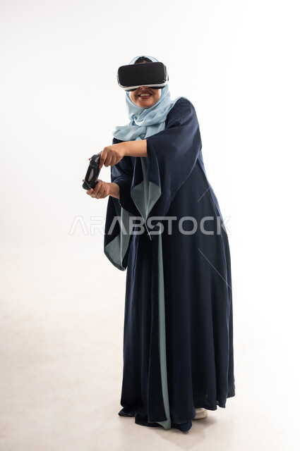 استخدام الأجهزة والتقنيات الحديثة، ايماءات الاستمتاع والاندماج، بورتريه لإمرأة عربية خليجية عمانية مبتسمة ترتدي الحجاب والعباءة تضع نظارة الواقع الإفتراضي، شابة سعودية تمسك بيدها عصا التحكم، اللعب بألعاب الفيديو الترفيهية، صورة الجسم بالكامل، خلفية بيضاء