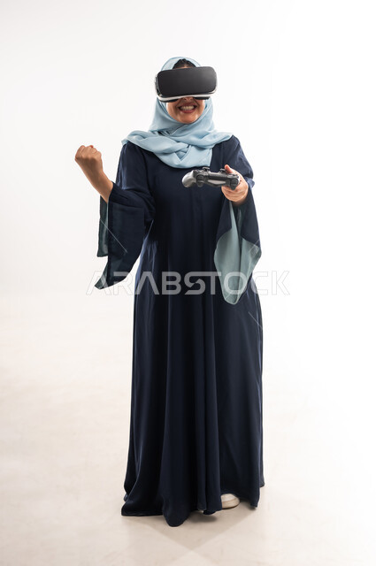 Playing recreational video games, a young Saudi woman playing with expressions of victory and winning, combining fun and entertainment with the imaginary world, portrait of a veiled Gulf Arab Omani woman wearing an abaya and virtual reality glasses holding a joystick in her hand, full body length, white background