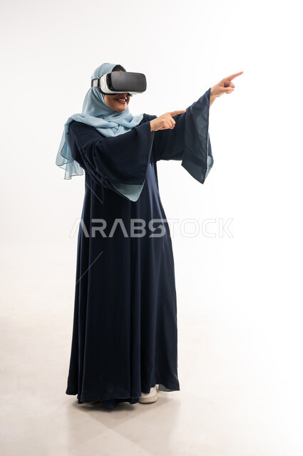 الاشارة الى مكان معين, المتعة  بالمشاهدة عبر نظارة الواقع الافتراضي, بورتريه لإمرأة عربية خليجية عمانية ترتدي العباءة تضع نظارات الفي آر, محاكاة العالم الخيالي, شابة سعودية تشير باصبعيها السبابة جانيا بتعابير هذا المكان هنا، صورة الجسم بالكامل، خلفية بيضاء