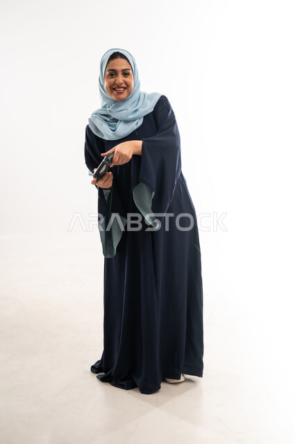 قضاء اوقات ممتعة بالعاب الفيديوز في عطلة نهاية الاسبوع، دمج التسلية بالتقنية والتكنولوجيا, بورتريه لإمرأة عربية خليجية عمانية محجبة ترتدي العباءة تحمل بيدها عصا التحكم بتعابير السعادة، شابة سعودية تلعب بإيماءات الاستمتاع، صورة الجسم بالكامل، خلفية بيضاء