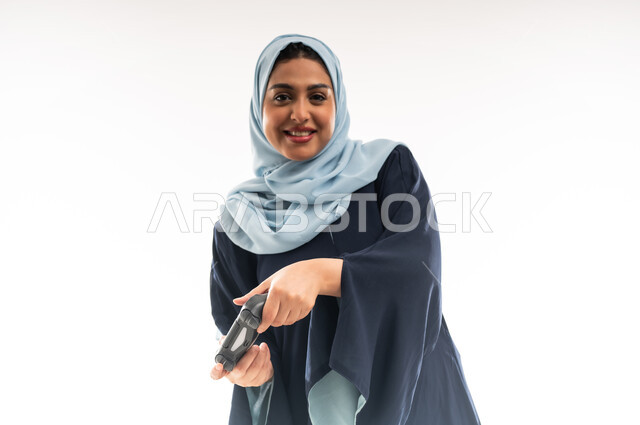 قضاء اوقات ممتعة بالعاب الفيديوز في عطلة نهاية الاسبوع، دمج التسلية بالتقنية والتكنولوجيا, شابة سعودية تلعب بإيماءات الاستمتاع والاندماج والتفاعل، بورتريه مقرب لإمرأة عربية خليجية عمانية محجبة ترتدي العباءة تحمل بيدها عصا التحكم بتعابير السعادة، خلفية بيضاء