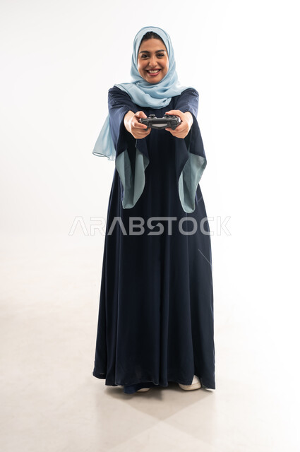 قضاء اوقات ممتعة بالعاب الفيديوز في عطلة نهاية الاسبوع، دمج التسلية بالتقنية والتكنولوجيا, بورتريه لإمرأة عربية خليجية عمانية محجبة ترتدي العباءة تحمل بيدها عصا التحكم بتعابير السعادة، شابة سعودية تلعب بإيماءات الاستمتاع، صورة الجسم بالكامل، خلفية بيضاء