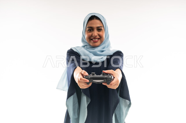 قضاء اوقات ممتعة بالعاب الفيديوز الترفيهية في عطلة نهاية الاسبوع، دمج التسلية بالتقنية والتكنولوجيا, شابة سعودية تلعب بإيماءات الاستمتاع والاندماج والتفاعل، بورتريه مقرب لإمرأة عربية خليجية عمانية محجبة ترتدي العباءة تحمل بيدها عصا التحكم بتعابير السعادة، خلفية بيضاء