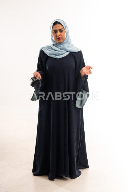 دمج التسلية  بالتكنولوجيا والتقنية الحديثة، العالم الرقمي الترفيهي، شابة سعودية تلعب بألعاب الفيديو الترفيهية, بورتريه لإمرأة عربية خليجية عمانية محجبة ترتدي العباءة تحمل عصا التحكم بتعابير الحزن والخسارة ، صورة الجسم بالكامل ،خلفية بيضاء