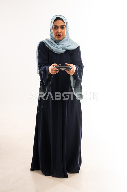 Fun and entertainment with video games, using modern electronic technologies, portrait of a veiled Gulf Omani Arab woman wearing an abaya holding a joystick in her hand, a young Saudi woman playing recreational games, gestures of enjoyment, integration and interaction, full body image, white background