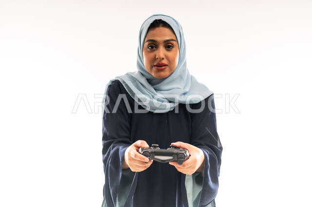 استخدام التقنيات الالكترونية الحديثة, بورتريه مقرب لإمرأة عربية خليجية عمانية محجبة ترتدي العباءة تحمل بيدها عصا التحكم، شابة سعودية تلعب بالألعاب الترفيهية، المتعة والتسلية بالفيديو جيمز في عطلة نهاية الاسبوع، إيماءات الاستمتاع والاندماج والتفاعل، خلفية بيضاء