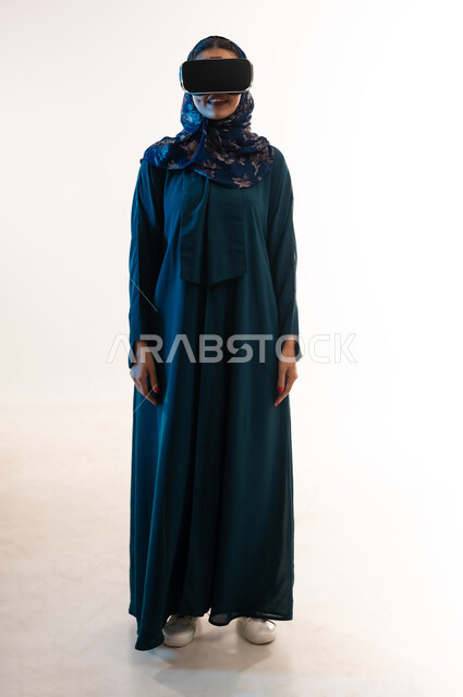 التسلية بالمشاهدة عبر نظارة الواقع الافتراضي،  بورتريه لإمرأة عربية خليجية عمانية محجبة ترتدي العباءة وتنظر من خلال نظارات ثلاثية الأبعاد، الاندماج ومحاكاة العالم الخيالي ، شابة سعودية تقف باستقامة، ايماءات السعادة والسرور, الطول الكامل للجسم، خلفيه يضاء