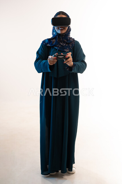 استخدام الأجهزة والتقنيات الحديثة، ايماءات الاستمتاع والاندماج والتفاعل، بورتريه لإمرأة عربية خليجية عمانية مبتسمة ترتدي الحجاب والعباءة تضع نظارة الواقع الإفتراضي وتمسك بيدها عصا التحكم، شابة سعودة تلعب بألعاب الفيديو الترفيهية، صورة الجسم بالكامل، خلفية بيضاء