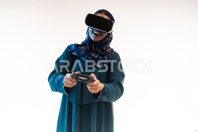 دمج التسلية مع العالم الخيالي، استخدام التقنية والتكنولوجيا، ايماءات الاستمتاع والاندماج والسعادة، بورتريه لإمرأة عربية خليجية عمانية محجبة ترتدي العباءة ونظارة الواقع الإفتراضي تمسك بيدها عصا التحكم، شابة سعودية تلعب بألعاب الفيديو الترفيهية، خلفية بيضاء
