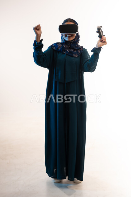 دمج المتعة والتسلية مع العالم الخيالي، شابة سعودية ترفع قبضة يدها بايماءات الفوز والانتصار، اللعب بألعاب الفيديو الترفيهية، بورتريه لإمرأة عربية خليجية عمانية محجبة ترتدي العباءة ونظارة الواقع الإفتراضي تمسك بيدها عصا التحكم، الطول الكامل للجسم، خلفية بيضاء