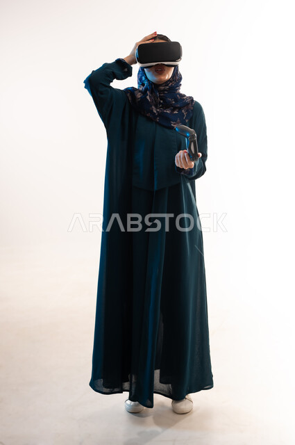 دمج التسلية بالتقنية والتكنولوجيا، محاكاة العالم الخيالي، شابة سعودية تضع يدها على رأسها بايماءات الدهشة والاستغراب، بورتريه لإمرأة عربية خليجية عمانية محجبة ترتدي العباءة تضع نظارة الواقع الافتراضي، التفاعل مع فيديو ثلاثي الأبعاد، الطول الكامل للجسم، خلفيه بيضاء