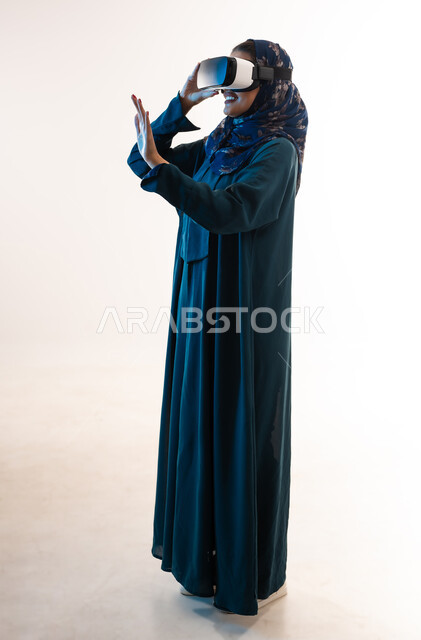 الاندماج ومحاكاة العالم الخيالي, شابة سعودية ترفع يدها لمحاولة لمس شيء، بورتريه من الجانب لإمرأة عربية خليجية عمانية ترتدي العباءة تضع نظارة ثلاثية الأبعاد تقوم بالتحرك والتفاعل معها، استخدام تقنية الواقع الافتراضي, صورة الجسم بالكامل، خلفية بيضاء