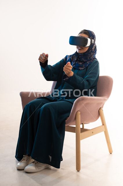 مفهوم استخدام التكنولوجيا والتقنيات الحديثة، الاندماج ومحاكاة العالم الخيالي، بورتريه لإمرأة عربية خليجية عمانية محجبة ترتدي العباءة وتضع نظارة الواقع الافتراضي، شابة سعودية تجلس على كرسي مريح تغلق قبضة يدها بشدة بايماءات النصر والقوة والتشجيع، خلفية بيضاء