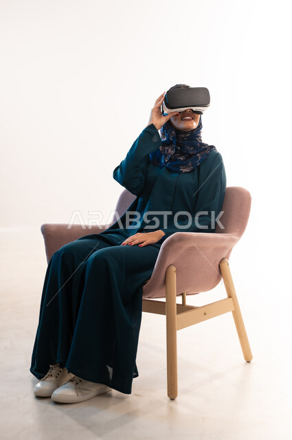 التطور التقني والتكنولوجي, المتعة و التسلية بالمشاهدة عبر نظارة الواقع الافتراضي،  بورتريه لإمرأة عربية خليجية عمانية ترتدي العباءة تنظر من خلال نظارات ثلاثية الأبعاد، الاندماج ومحاكاة العالم الخيالي ،شابة سعودية تجلس على كرسي مريح بايماءات السعادة، خلفيه يضاء