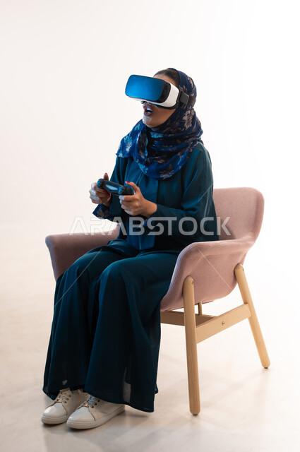 استخدام الأجهزة الحديثة، دمج التسلية مع العالم الخيالي، بورتريه لإمرأة عربية خليجية عمانية محجبة ترتدي العباءة ونظارة الواقع الإفتراضي تجلس على كرسي مريح تمسك بيدها عصا التحكم بتعابير الاندهاش والذهول، شابة سعودية تلعب بألعاب الفيديو الترفيهية، خلفية بيضاء