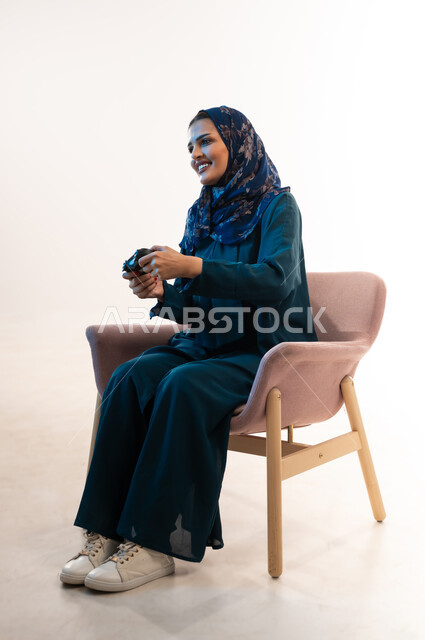 دمج التسلية بالتقنية والتكنولوجيا، شابة سعودية مبتسمة تلعب بايماءات الاندماج والتفاعل، قضاء اوقات ممتعة  بألعاب الفيديوز الترفيهية في عطلة نهاية الاسبوع ، بورتريه لإمرأة عربية خليجية عمانية ترتدي العباءة تجلس على كرسي مريح تحمل بيدها عصا التحكم، خلفية بيضاء