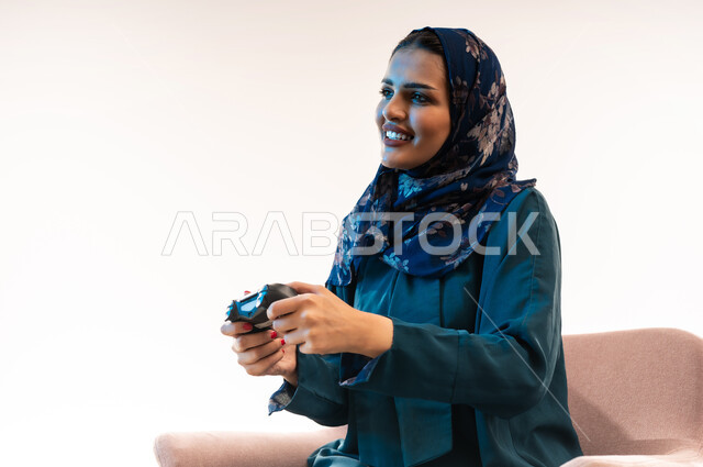 دمج التسلية بالتقنية والتكنولوجيا، شابة سعودية مبتسمة تلعب بايماءات الاندماج والتفاعل، قضاء اوقات ممتعة  بألعاب الفيديوز الترفيهية في عطلة نهاية الاسبوع ، بورتريه مقرب لإمرأة عربية خليجية عمانية ترتدي العباءة تجلس على كرسي مريح تحمل بيدها عصا التحكم، خلفية بيضاء