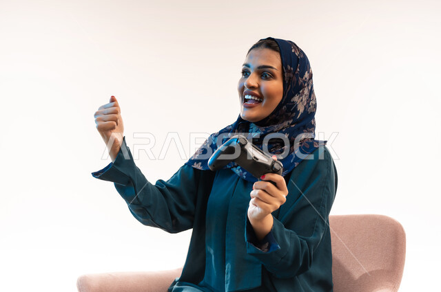 استخدام التقنية والتكنولوجيا، قضاء اوقات ممتعة  بألعاب الفيديوز في عطلة نهاية الاسبوع، بورتريه مقرب لإمرأة عربية خليجية عمانية ترتدي العباءة تجلس على كرسي مريح تحمل بيدها عصا التحكم، شابة سعودية مبتسمة تغلق قبضة اليد بتعابير الفوز والنصر والقوة، خلفية بيضاء