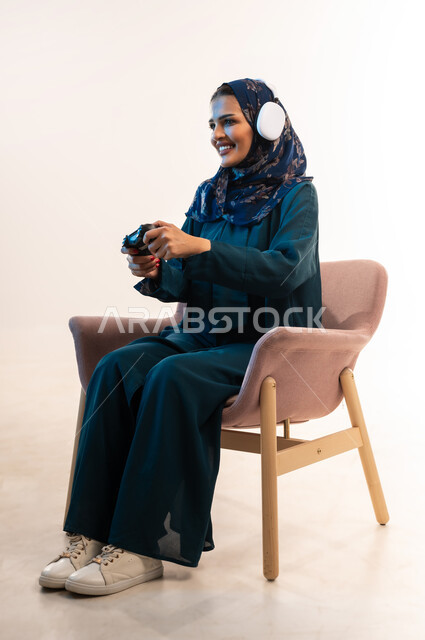 Having fun playing video games, gestures of integration while playing, using modern and advanced electronic technologies, portrait of an Arab Gulf Omani woman wearing a headset holding a joystick in her hand, a young Saudi woman sitting on a comfortable chair enjoying her vacation, white background