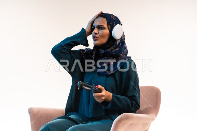 استخدام التقنيات الحديثة، تعابير الاحباط للخسارة، قضاء اوقات ممتعة بلعب ألعاب الفيديو الترفيهية، شابة سعودية تضع يدها على راسها بايماءات الحزن، بورتريه مقرب من الجانب لإمرأة عربية خليجية عمانية ترتدي سماعة الرأس تجلس على كرسي مريح تحمل بيدها عصا التحكم، خلفية بيضاء