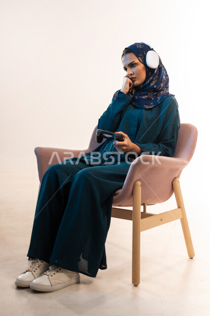 قضاء اوقات ممتعة بألعاب الفيديوز، استخدام التقنيات الالكترونية الحديثة والمتطورة، بورتريه من الجانب لامرأة عربية خليجية عمانية ترتدي سماعة الرأس تجلس على كرسي مريح تحمل بيدها عصا التحكم، شابة سعودية ضع يدها على ذقنها بتعابير الشرود، خلفية بيضاء