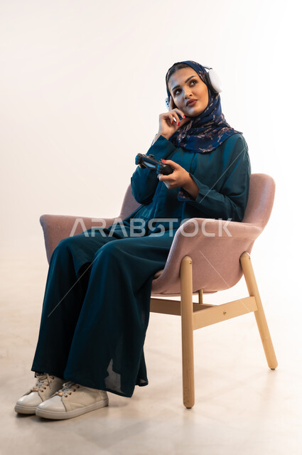 Having fun playing video games, using modern and advanced electronic technologies, side portrait of an Arab Gulf Omani woman wearing headphones sitting on a comfortable chair holding a joystick in her hand, a young Saudi woman putting her hand on her cheek with a thinking expression, white background