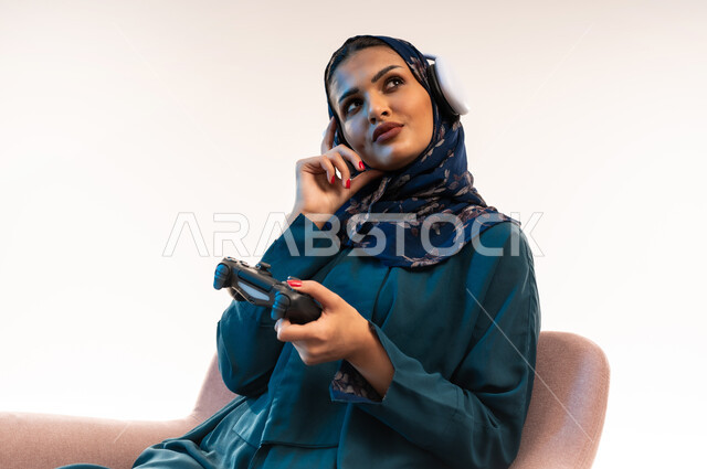 استخدام التقنيات الالكترونية الحديثة والمتطورة، قضاء اوقات ممتعة بألعاب الفيديوز، بورتريه مقرب من الجانب لامرأة عربية خليجية عمانية ترتدي سماعة الرأس تجلس على كرسي مريح تحمل بيدها عصا التحكم، شابة سعودية ضع يدها على خدها بتعابير التفكير، خلفية بيضاء