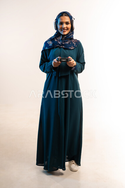 قضاء اوقات ممتعة بألعاب الفيديوز، ايماءات الاندماج أثناء اللعب، استخدام التقنيات الالكترونية الحديثة والمتطورة، بورتريه لامرأة عربية خليجية عمانية ترتدي سماعة الرأس تحمل بيدها عصا التحكم، شابة سعودية تستمتع بالاجازة، صورة لكامل الجسم، خلفية بيضاء