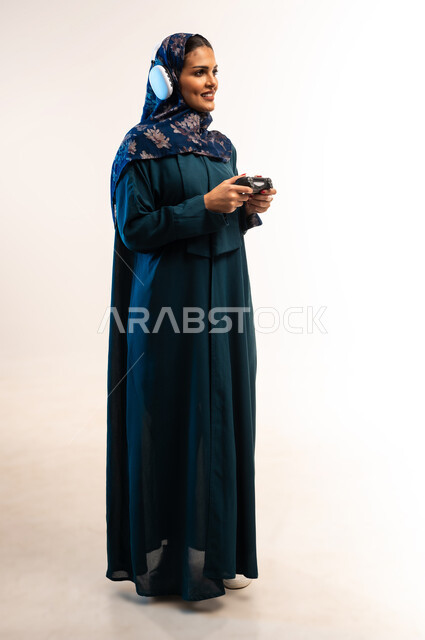 قضاء اوقات ممتعة بألعاب الفيديوز، ايماءات الاندماج أثناء اللعب، استخدام التقنيات الالكترونية الحديثة والمتطورة، بورتريه من الجانب لامرأة عربية خليجية عمانية ترتدي سماعة الرأس تحمل بيدها عصا التحكم، شابة سعودية تستمتع بالاجازة، صورة لكامل الجسم، خلفية بيضاء