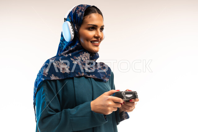 ايماءات الاندماج والتفاعل أثناء اللعب، قضاء اوقات ممتعة بألعاب الفيديوز الترفيهية، استخدام التقنيات الالكترونية الحديثة والمتطورة، بورتريه مقرب من الجانب لامرأة عربية خليجية عمانية ترتدي سماعة الرأس تحمل بيدها عصا التحكم، شابة سعودية تستمتع بالاجازة، خلفية بيضاء