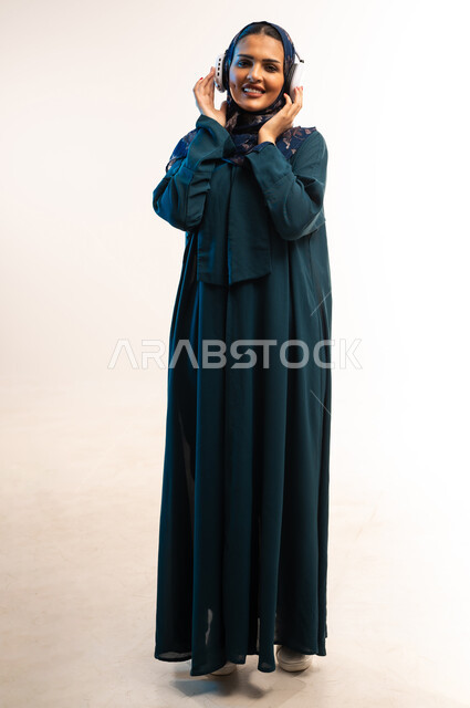 قضاء اوقات ممتعة ومسلية، مفهوم الإصغاء والإستماع، إستخدام تقنيات الهيدفون، بورتريه لامرأة عربية خليجية عمانية محجبة تضع سماعات الرأس الصوتية تستمع إلى الموسيقى، شابة سعودية تنظر الكاميرا بتعابير السعادة والسرور، صورة لكامل الجسم، خلفية بيضاء