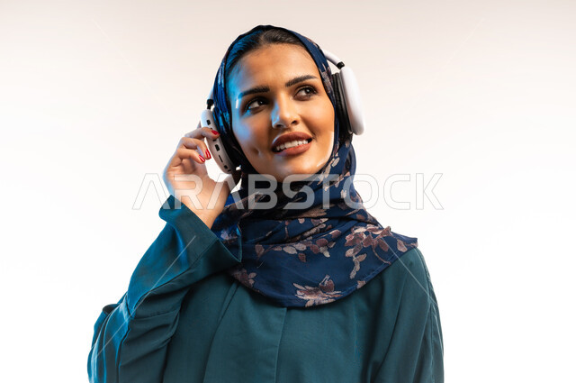 استخدام تقنيات الهيدفون، مفهوم الإصغاء والإستماع، قضاء اوقات ممتعة ومسلية، بورتريه مقرب لامرأة عربية خليجية عمانية محجبة تضع سماعات الرأس الصوتية تستمع إلى الموسيقى، شابة سعودية تنظر لمكان ما بتعابير السعادة والسرور، خلفية بيضاء