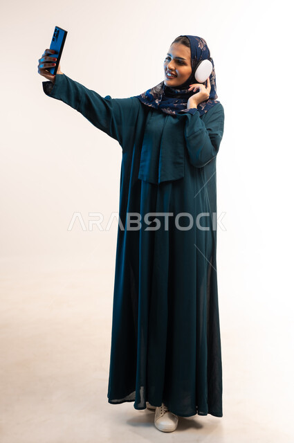 التقاط صور السيلفي التذكارية، قضاء اوقات ممتعة في عطلة نهاية الاسبوع، إستخدام تقنيات الهيدفون، بورتريه لامرأة عربية خليجية عمانية محجبة ترتدي العباءة تضع سماعة الرأس تقوم بإجراء مكالمة فيديو، شابة سعودية تستخدام الهاتف لمحمول، الجسم الكامل بالطول، خلفية بيضاء