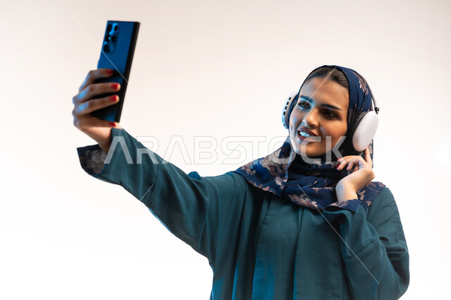 قضاء اوقات ممتعة في عطلة نهاية الاسبوع، التقاط صور السيلفي التذكارية، إستخدام تقنيات الهيدفون، بورتريه مقرب لامرأة عربية خليجية عمانية محجبة ترتدي العباءة تضع سماعة الرأس تقوم بإجراء مكالمة فيديو، شابة سعودية تستخدام الهاتف لمحمول، خلفية بيضاء