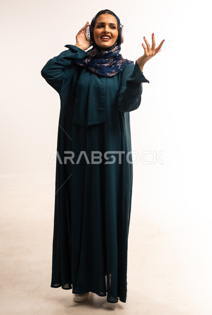 Concept of listening, using headphones, having fun and entertaining time, portrait of a veiled Omani Gulf Arab woman wearing headphones listening to music, young Saudi woman raising her hand with gestures of integration, full body image, white background