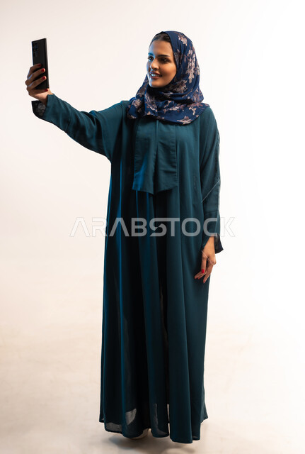 التقاط صور السيلفي التذكارية، قضاء اوقات ممتعة في عطلة نهاية الاسبوع، استخدام التقنيات الحديثة، بورتريه لامرأة عربية خليجية عمانية محجبة ترتدي عباءة زرقاء تقوم بإجراء مكالمة فيديو، شابة سعودية تستخدام الهاتف لمحمول، صورة لكامل الجسم، خلفية بيضاء