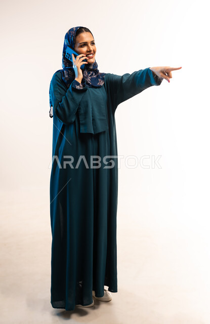 التواصل مع الأهل والأصدقاء، إجراء مكالمة صوتية عبر الجوال، دمج التقنيات والتكنولوجيا الحديثة في الحياة اليومية، بورتريه من الجانب لامرأة عربية خليجية عمانية ترتدي العباءة تشير بإصبع السبابة الى مكان ما، شابة تنظر تحمل بيدها الهاتف المحمول، الجسم الكامل بالطول، خلفية بيضاء