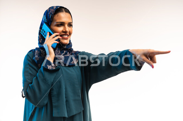 إجراء مكالمة صوتية عبر الجوال، التواصل مع الأهل والأصدقاء، دمج التقنيات والتكنولوجيا الحديثة في الحياة اليومية، بورتريه مقرب من الجانب لامرأة عربية خليجية عمانية ترتدي العباءة تشير بإصبع السبابة الى مكان ما، شابة تنظر تحمل بيدها الهاتف المحمول، خلفية بيضاء