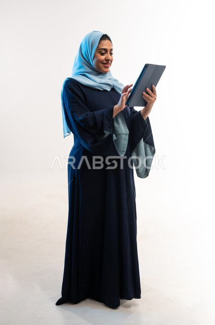 دمج التقنية والتكنولوجيا الحديثة في الحياة اليومية، بورتريه من الجانب لامرأة عربية خليجية عمانية ترتدي العباءة تقوم بالنقر على التابلت، تصفح مواقع ووسائل التواصل الاجتماعي، شابة سعودية تعمل على الجهاز اللوحي، صورة لكامل الجسم، خلفية بيضاء