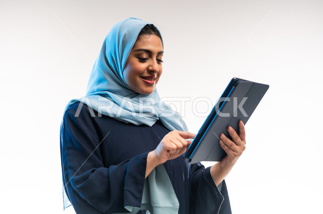 Browsing social media sites and media, integrating technology and modern technology into daily life, close-up side portrait of an Omani Gulf Arab woman wearing an abaya tapping on a tablet, young Saudi woman working on a tablet, white background