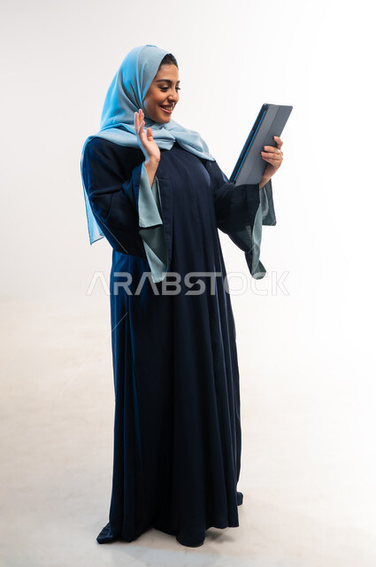 إلقاء التحية والترحيب والسلام، دمج التكنولوجيا الحديثة في الحياة اليومية، بورتريه لامرأة عربية خليجية عمانية ترتدي العباءة تقوم بإجراء مكالمة فيديو، شابة سعودية تستخدام جهاز التابلت، التواصل مع الأهل والأصدقاء، صورة لكامل الجسم، خلفية بيضاء