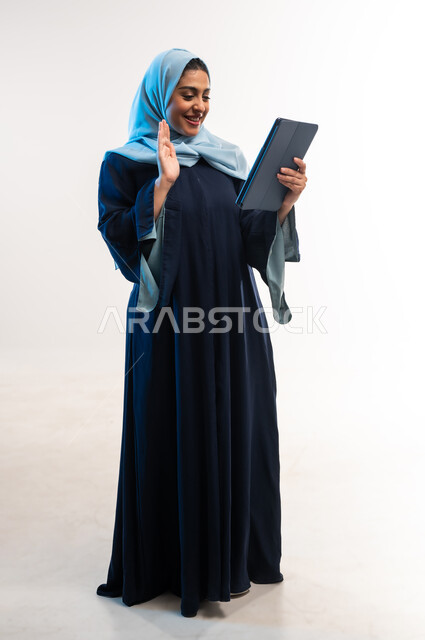 التواصل مع الأهل والأصدقاء، إلقاء التحية والترحيب والسلام، دمج التكنولوجيا الحديثة في الحياة اليومية، بورتريه لامرأة عربية خليجية عمانية ترتدي العباءة تقوم بإجراء مكالمة فيديو، شابة سعودية تستخدام جهاز التابلت، صورة لكامل الجسم، خلفية بيضاء