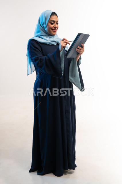 دمج التقنية والتكنولوجيا الحديثة في الحياة اليومية، تصفح مواقع ووسائل التواصل الاجتماعي، بورتري مقرب لامرأة عربية خليجية عمانية ترتدي العباءة تقوم بالكتابة على التابلت، مفهوم انجار المشاريع عن بعد، شابة سعودية تحمل بيدها الجهاز اللوحي، صورة لكامل الجسم، خلفية بيضاء