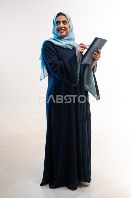 دمج التقنية والتكنولوجيا الحديثة في الحياة اليومية، تصفح مواقع ووسائل التواصل الاجتماعي، بورتري مقرب لامرأة عربية خليجية عمانية ترتدي العباءة تقوم بالكتابة على التابلت، مفهوم انجار المشاريع عن بعد، شابة سعودية تحمل بيدها الجهاز اللوحي، صورة لكامل الجسم، خلفية بيضاء