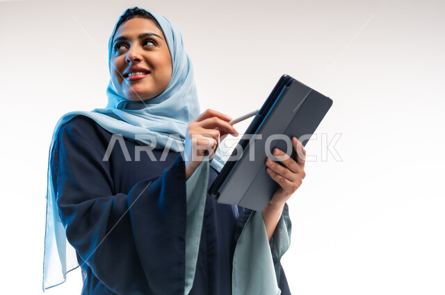 تصفح مواقع ووسائل التواصل الاجتماعي، دمج التقنية والتكنولوجيا الحديثة في الحياة اليومية والعملية، بورتريه مقرب لامرأة عربية خليجية عمانية ترتدي العباءة تقوم بالكتابة على التابلت، مفهوم انجار المشاريع عن بعد، شابة سعودية تحمل بيدها الجهاز اللوحي، خلفية بيضاء