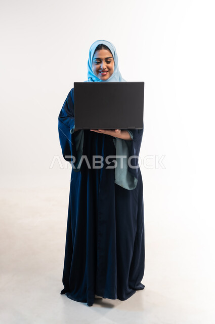 تصفح مواقع ووسائل التواصل الاجتماعي، دمج التقنية والتكنولوجيا الحديثة في الحياة اليومية، بورتريه لامرأة عربية خليجية عمانية ترتدي العباءة تقوم بالنقر على لوحة مفاتيح اللابتوب، شابة سعودية تعمل على جهاز الحاسوب المحمول، صورة لكامل الجسم، خلفية بيضاء