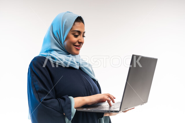 تعابير السرور والسعادة، تصفح مواقع ووسائل التواصل الاجتماعي، دمج التقنية والتكنولوجيا الحديثة في الحياة اليومية، بورتريه مقرب من الجانب لامرأة عربية خليجية عمانية تقوم بالنقر على لوحة مفاتيح اللابتوب، شابة سعودية تعمل على جهاز الحاسوب المحمول، خلفية بيضاء