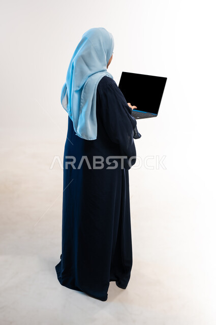 دمج التقنية والتكنولوجيا الحديثة في الحياة اليومية، بورتريه من الخلف لامرأة عربية خليجية عمانية ترتدي العباءة تقوم بالنقر على لوحة مفاتيح اللابتوب، تصفح مواقع ووسائل التواصل الاجتماعي، شابة سعودية تعمل على جهاز الحاسوب المحمول، صورة لكامل الجسم، خلفية بيضاء