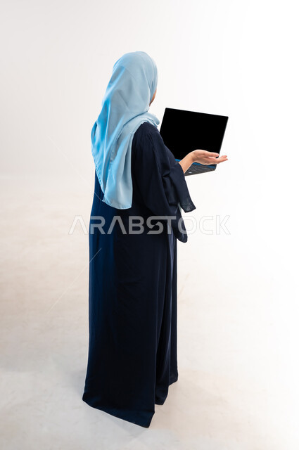 عرض شاشة الحاسوب المحمول السوداء، اجراء مقابلات عمل اونلاين، دمج التكنولوجيا الحديثة في الحياة العملية، بورتريه من الخلف لامرأة عربية خليجية عمانية ترتدي العباءة تحمل بيدها عنصر ما، شابة سعودية ترفع كفها بإيماءات عدم المعرفة، صورة لكامل الجسم، خلفية بيضاء